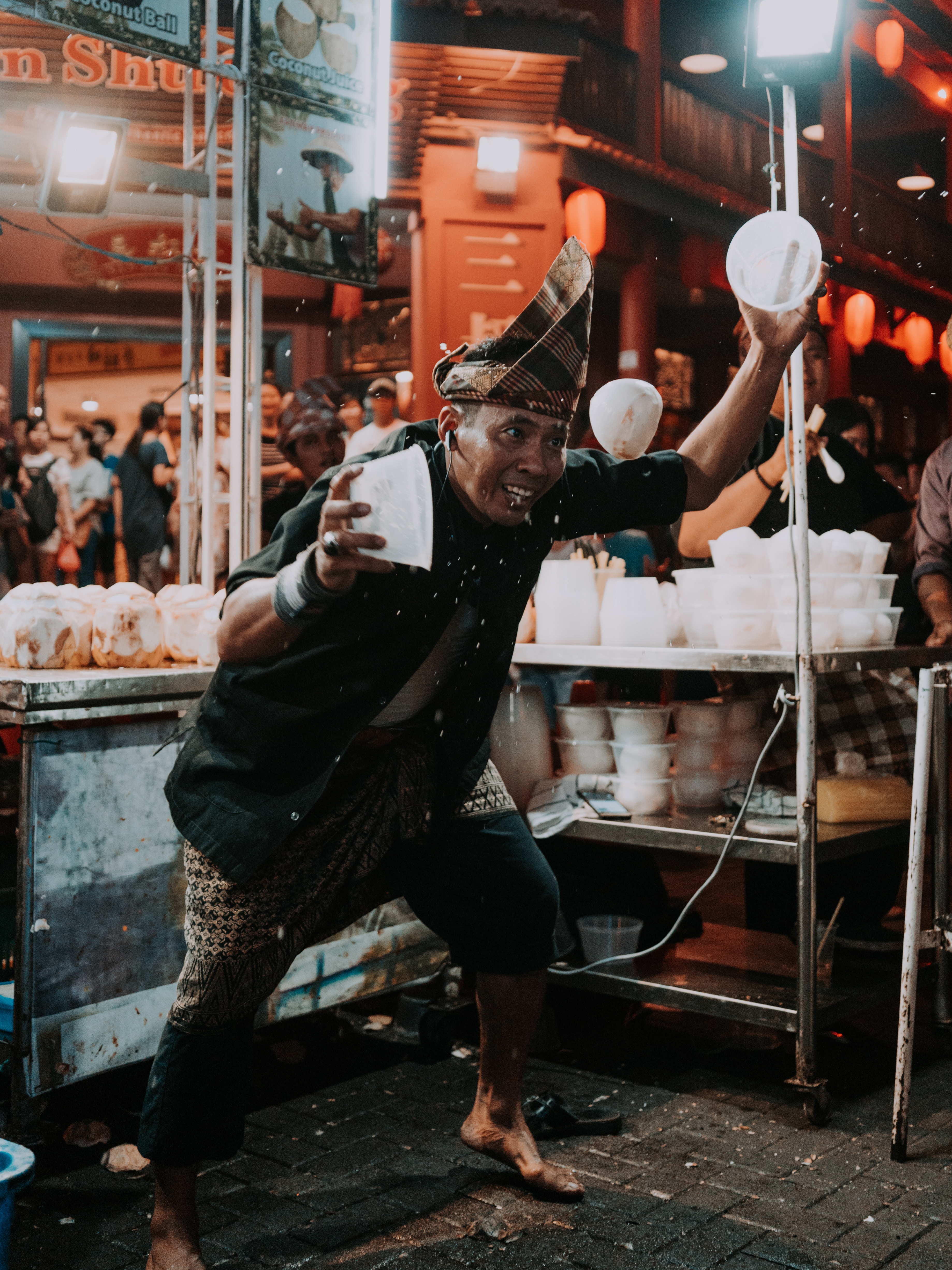 A barefoot street food vendor wearing a pointed hat and juggling ice between two bowls.