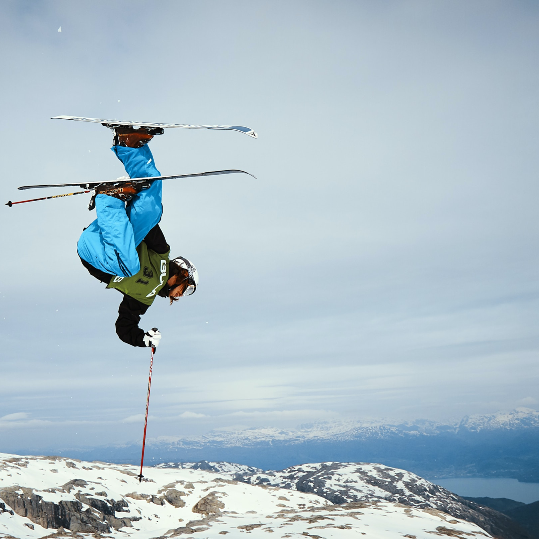 A skier doing a flip with his feet in the air above his head, on high, rocky terrain that overlooks a large body of water.