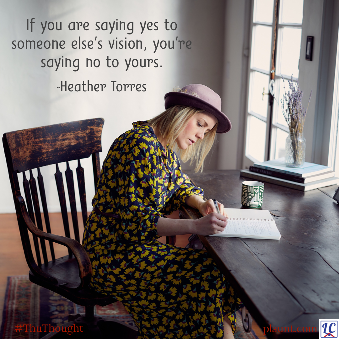 A woman in a floral print dress, wearing a hat, sitting at a wood plank table, writing in a notebook. Caption: If you are saying yes to someone else’s vision, you’re saying no to yours. ~Heather Torres