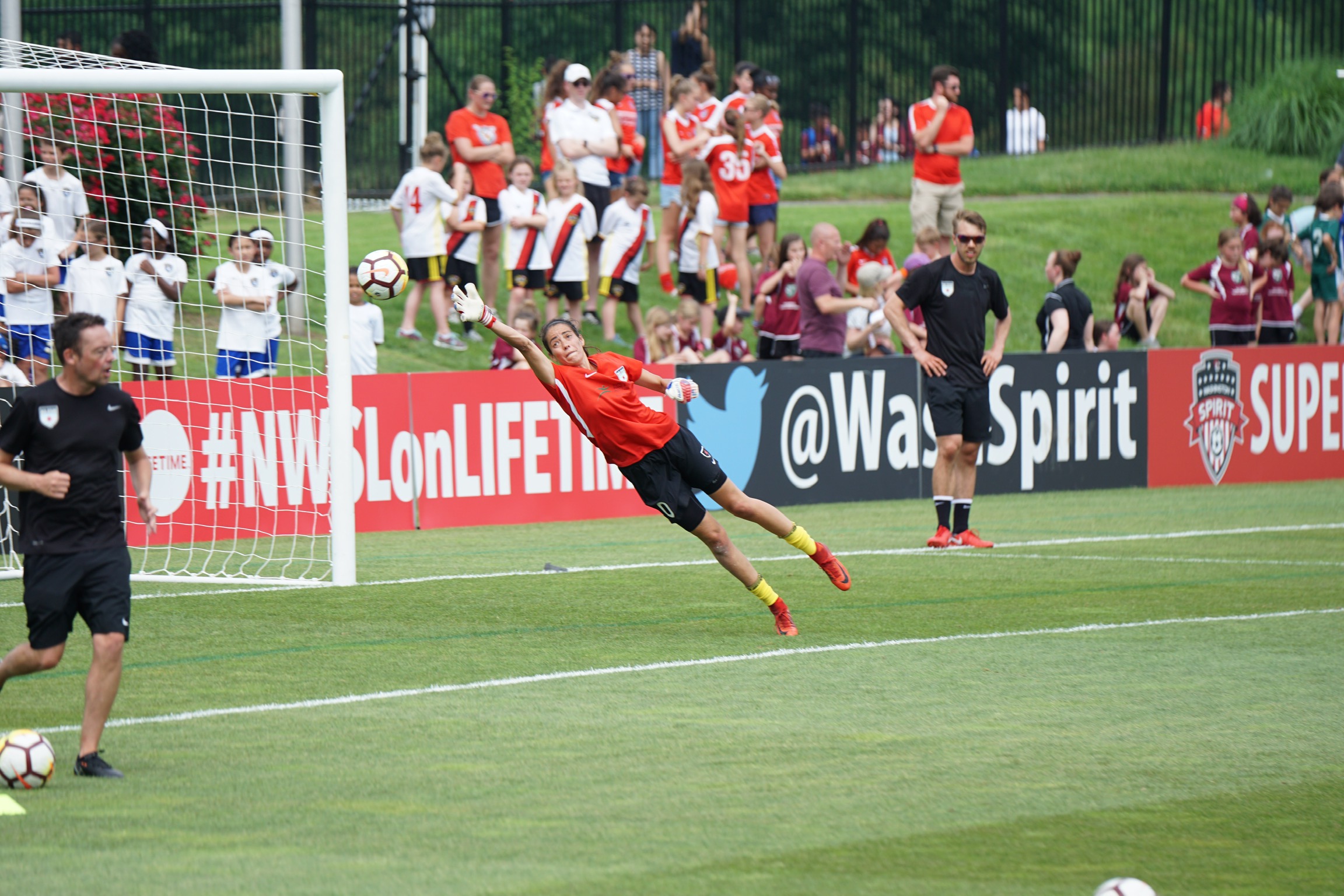 A soccer player elevated at a 45 degree angle from the ground, reaching for a soccer ball in front of the net and a crowd of onlookers on the sidelines.