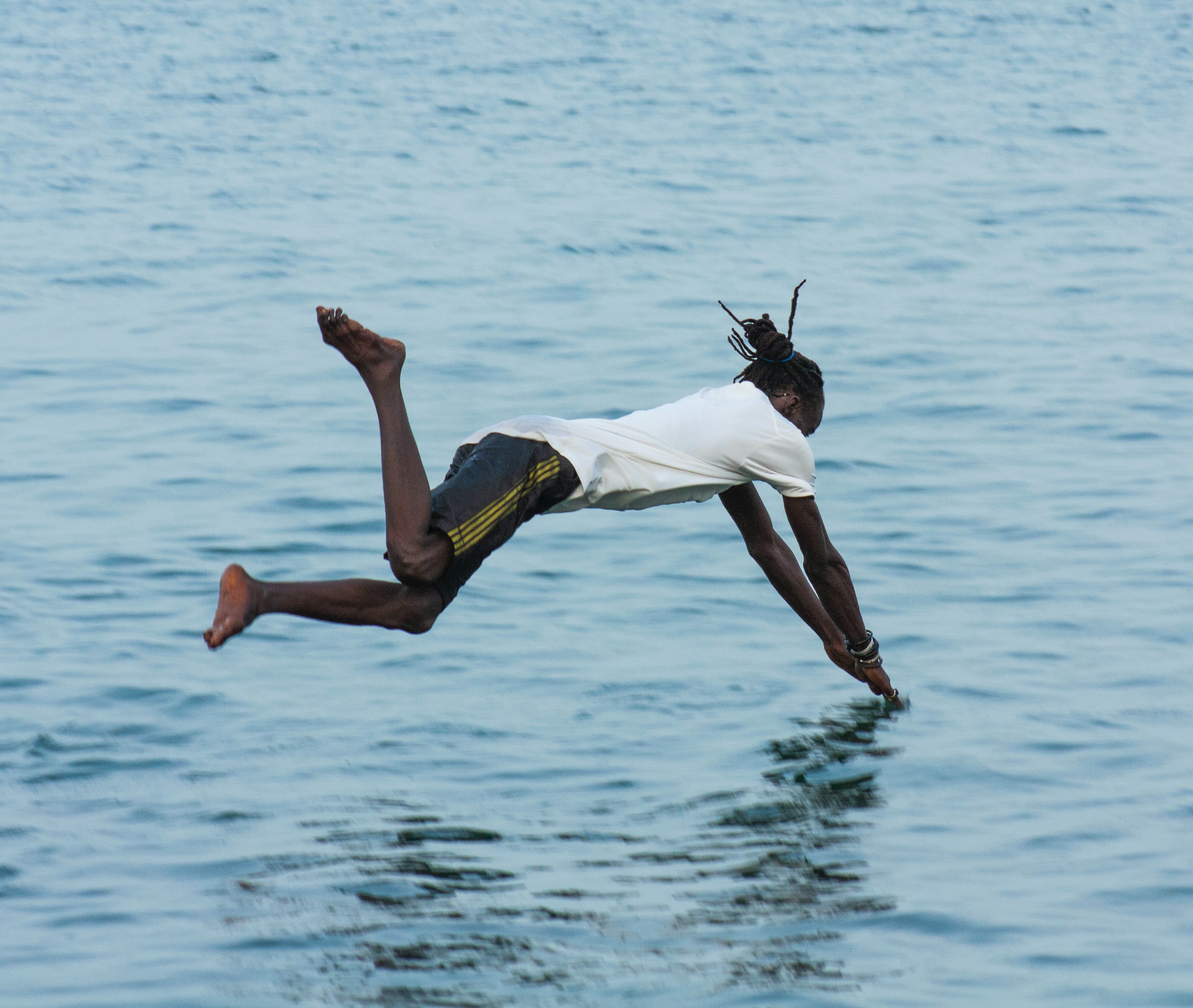 A man diving into, and captured just above, a body of water. His arms are straight, but his legs are bent at different angles. He may do a bellyflop.