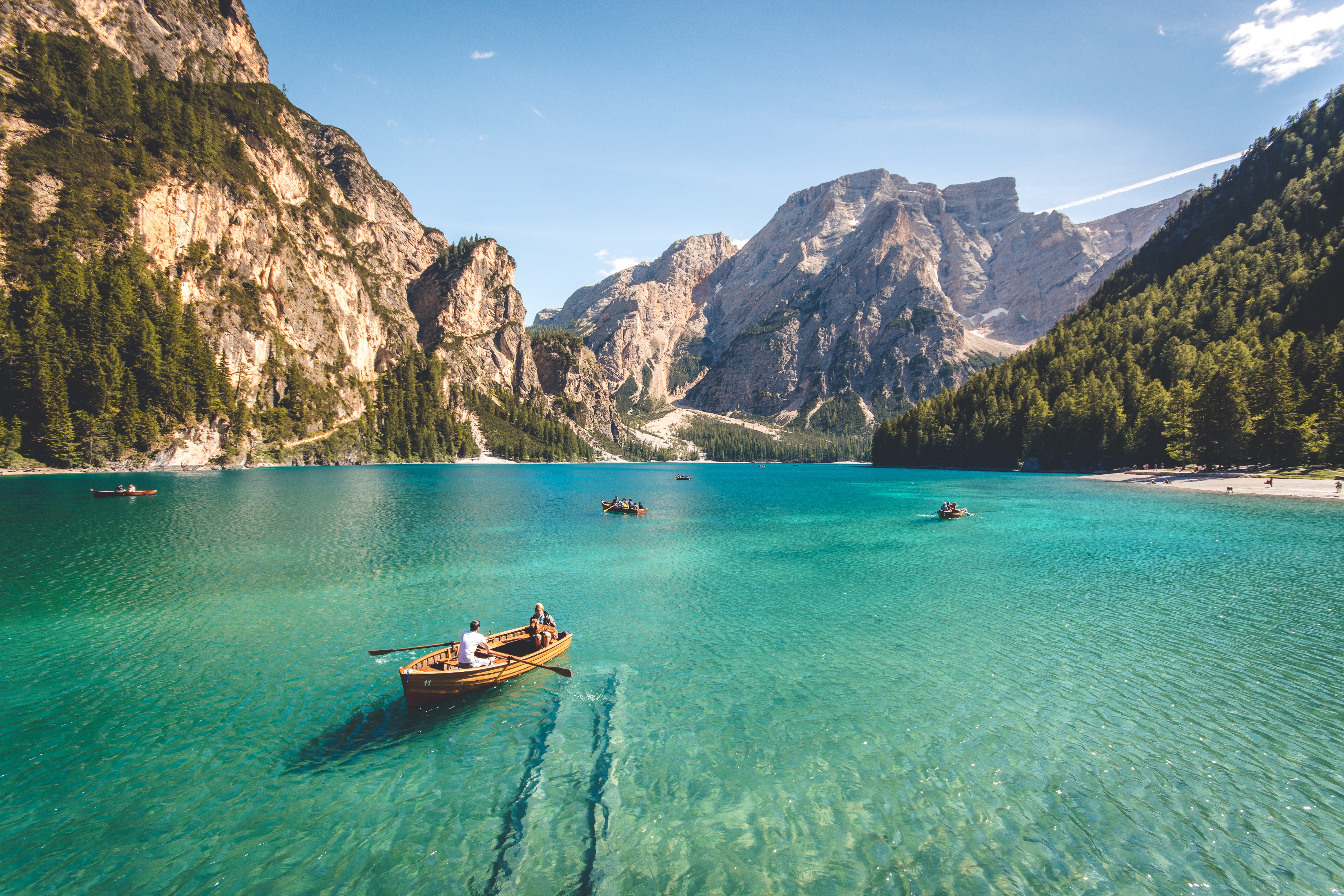 Several rowboats, with varying numbers of people in each, floating on a calm, aquamarine lake surrounded by mountains. The boat in the foreground has a man rowing and a woman sitting holding a dog.