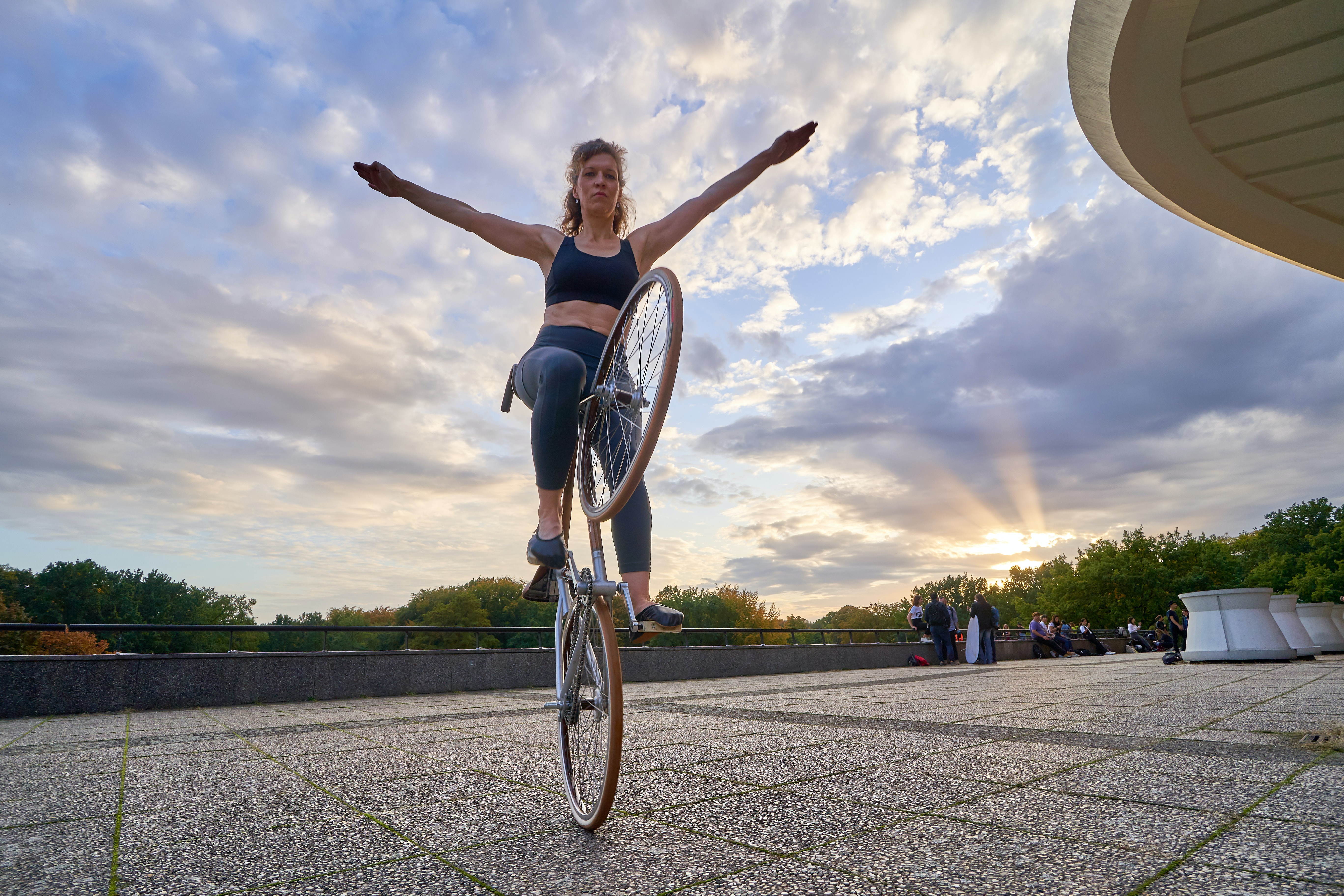 A woman with outstretched arms balancing on the back tire of a bicycle in a public square. The setting sun behind her is causing crepuscular rays in a cloudy sky.
