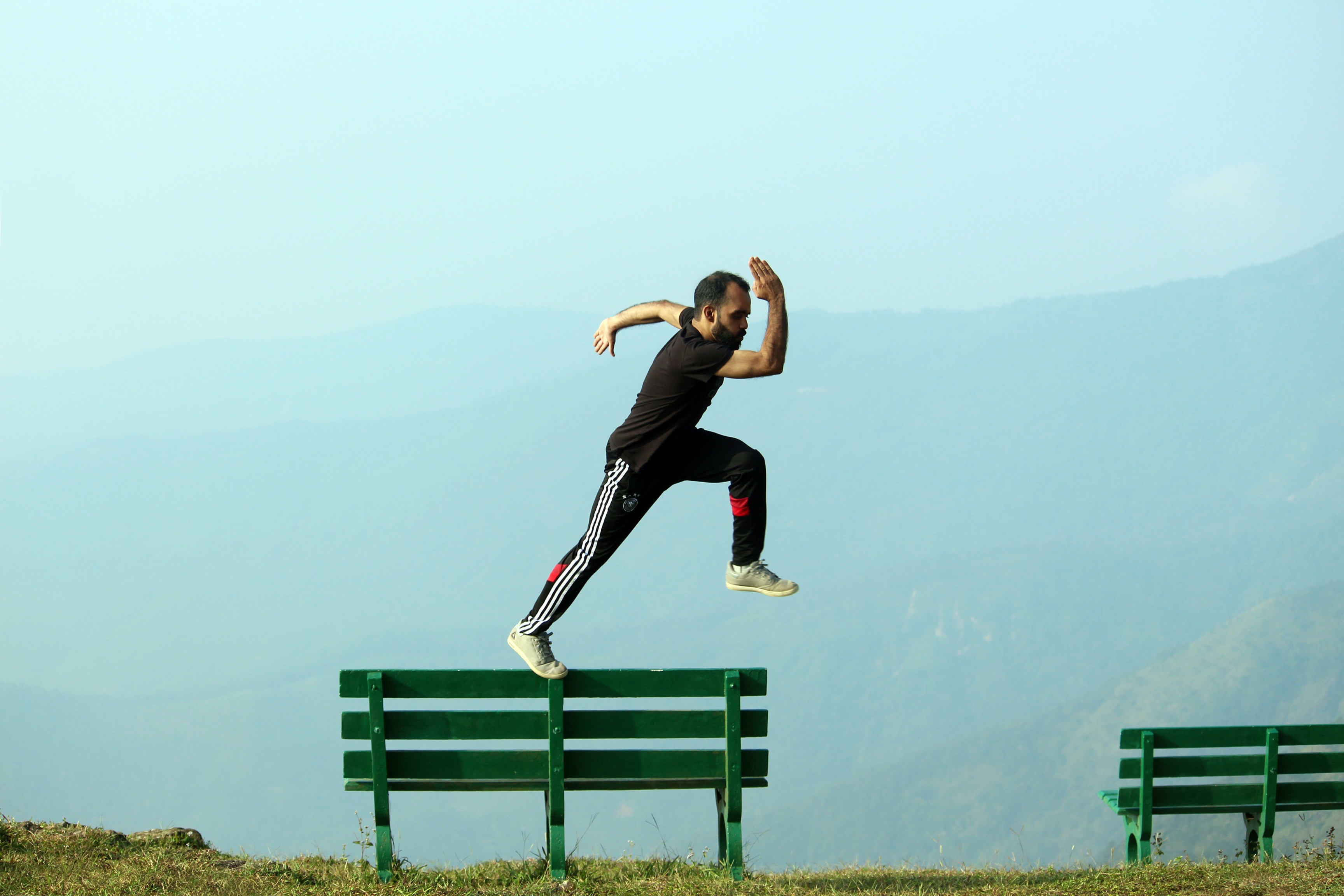 A man running across the back of a park bench looking like he is about to leap into the air. In the background is a mountain shrouded with mist with a deep valley below.