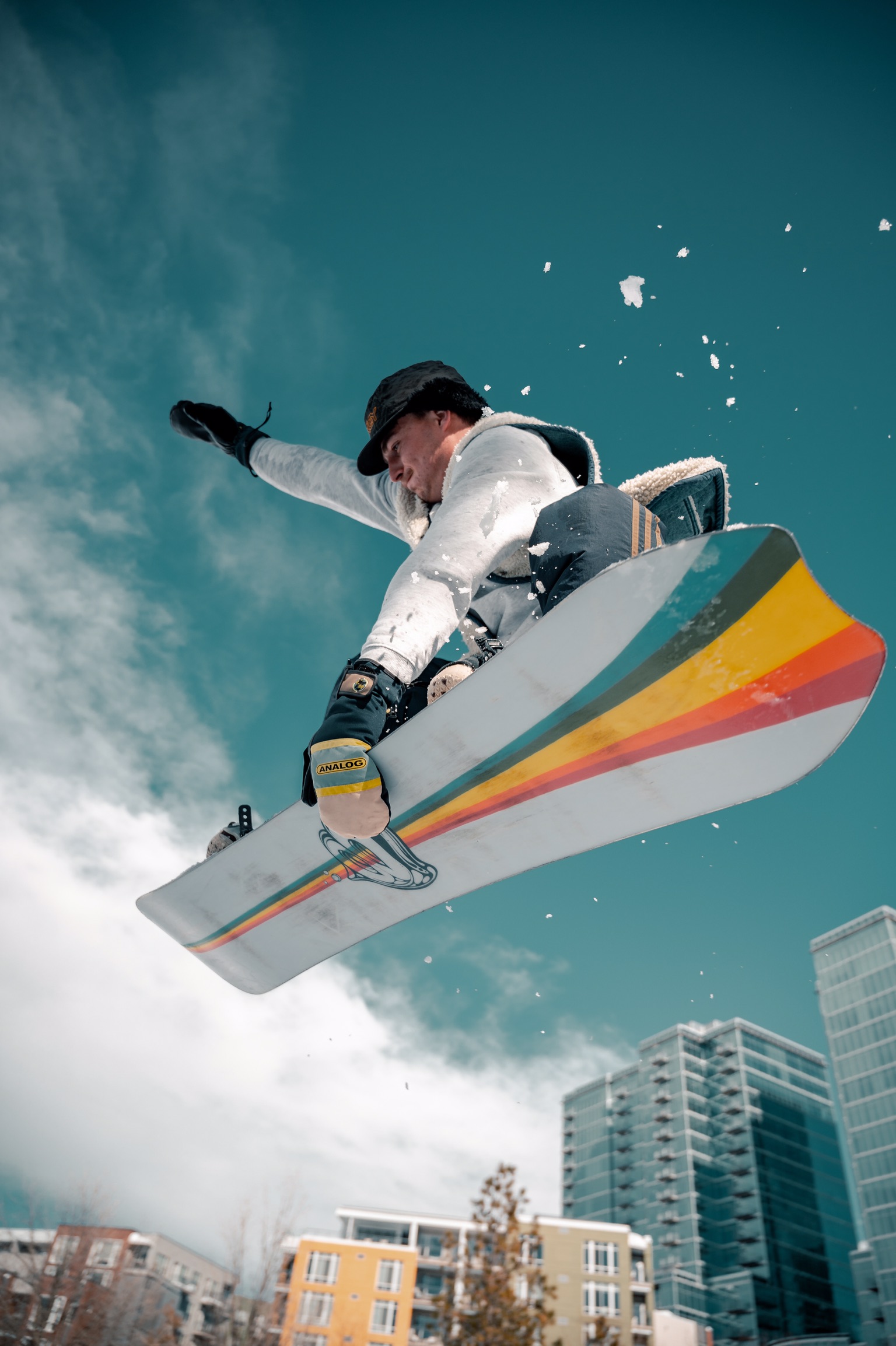 A person flying through the air on a white snowboard with multi-coloured stripes. There are tall city buildings in the background. A snow hill is not visible.