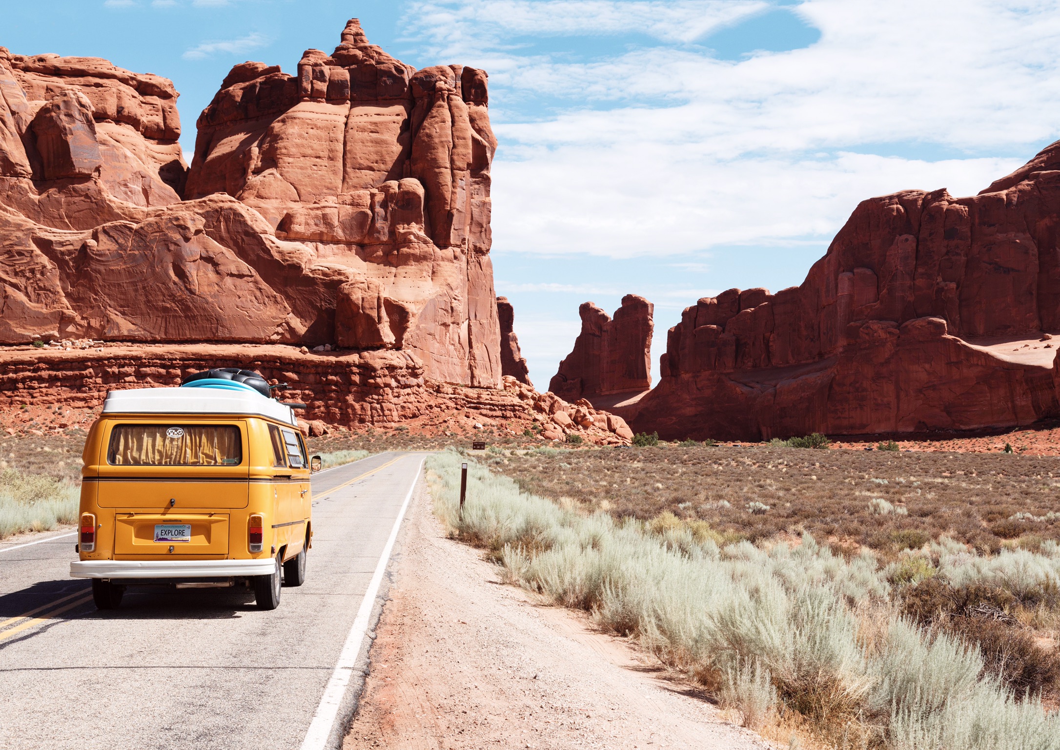 A mustard-coloured Volkswagen van on a road in front of the tall red rock formations in Arches National Park.