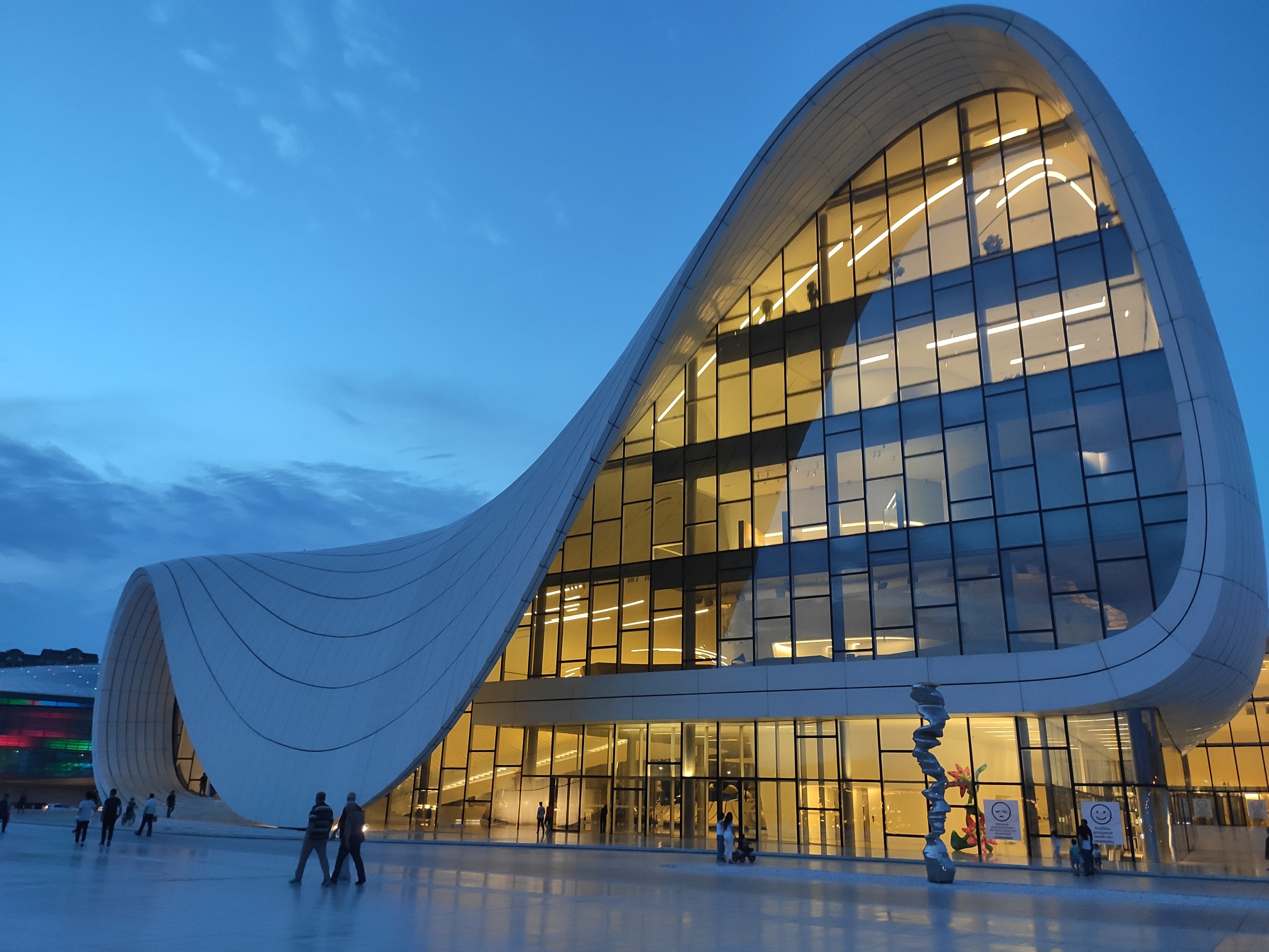 A modern white and glass building with a curved roof that appears to be melting on one side. Lights inside give a golden glow behind the glass.