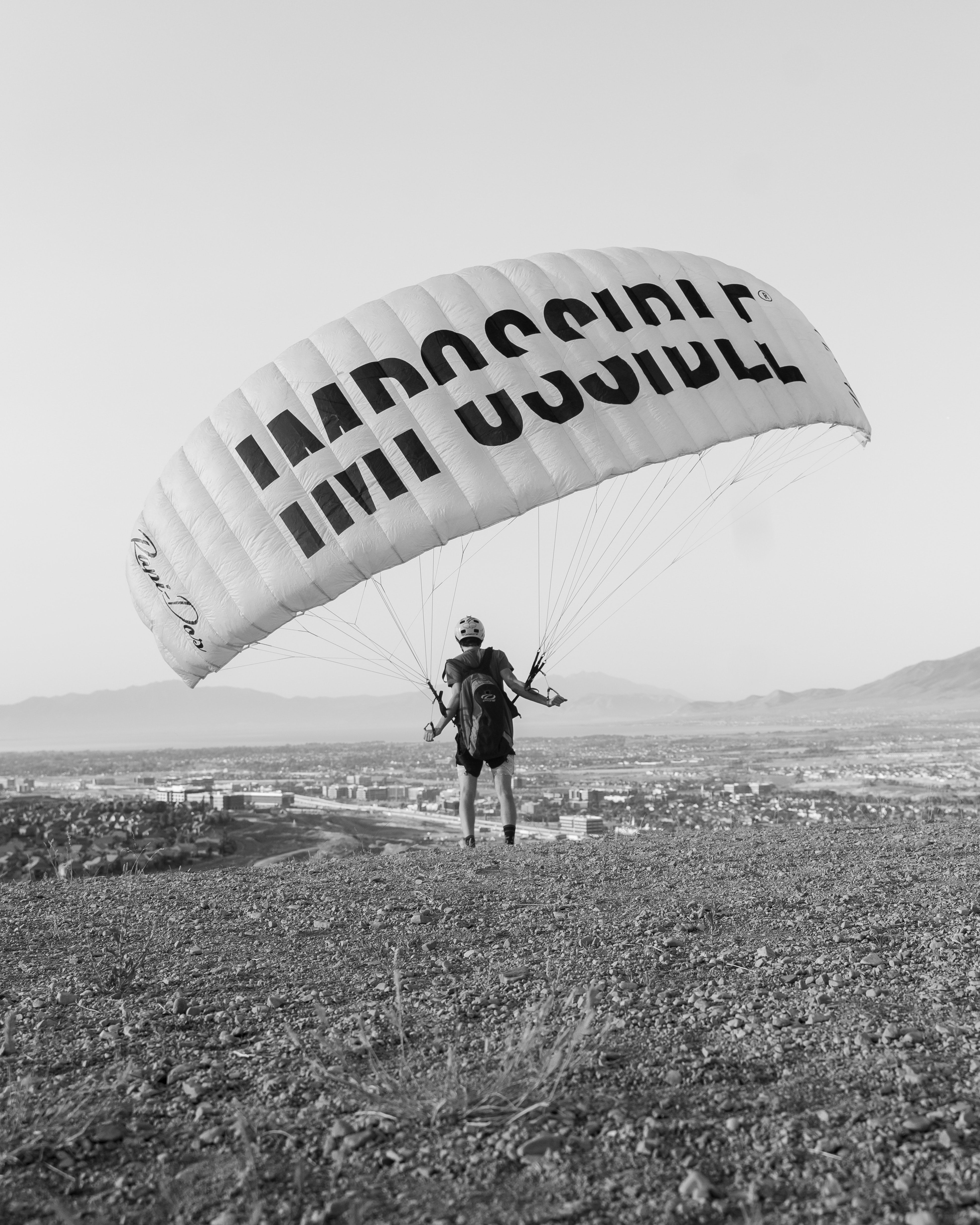 A person at the edge of a cliff prepared to base jump with a white parachute that says IMPOSSIBLE in big black letters with a white line through the middle.
