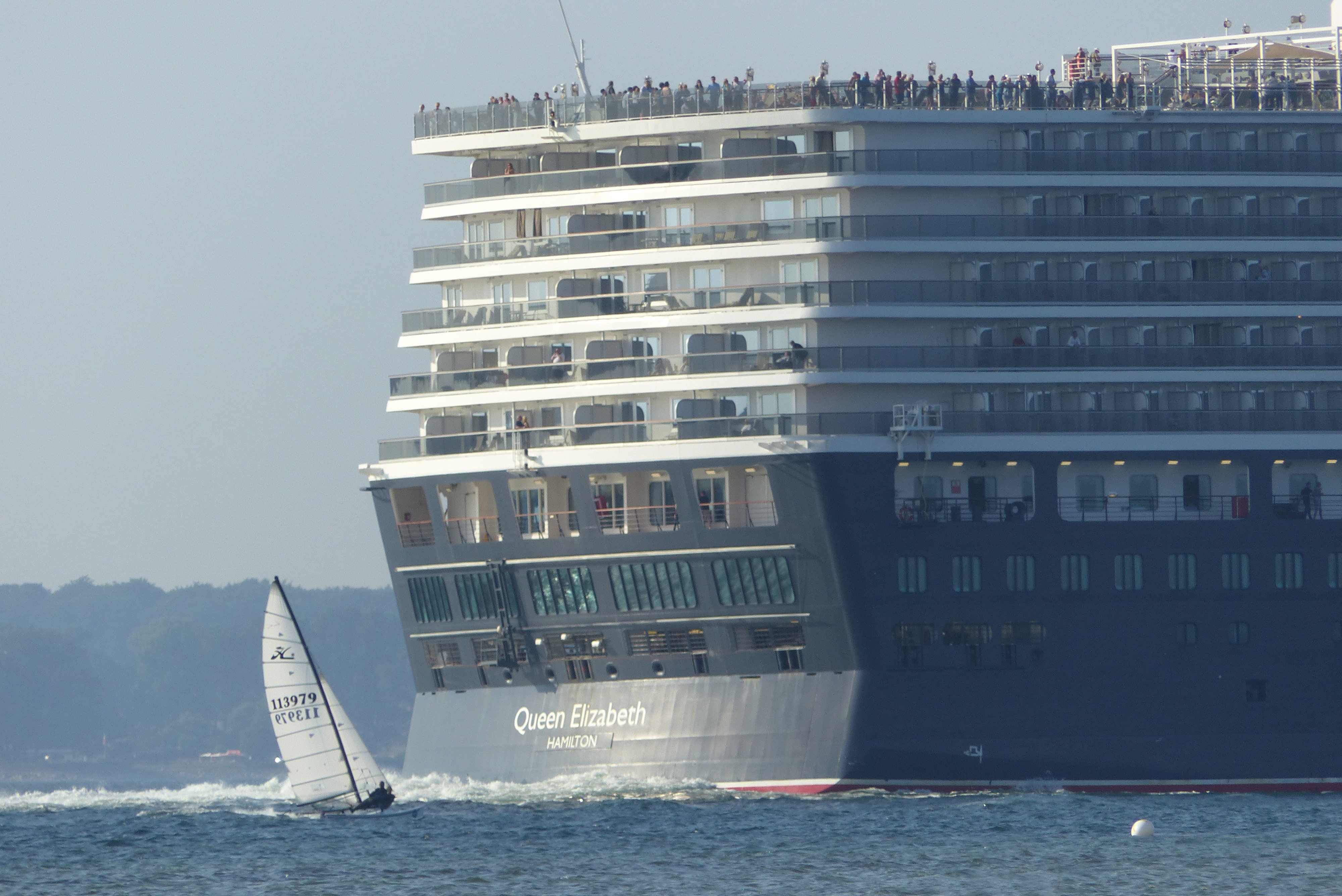 The Queen Elizabeth, a cruise ship the size of an apartment building, is at sea with a small sailboat beside it.