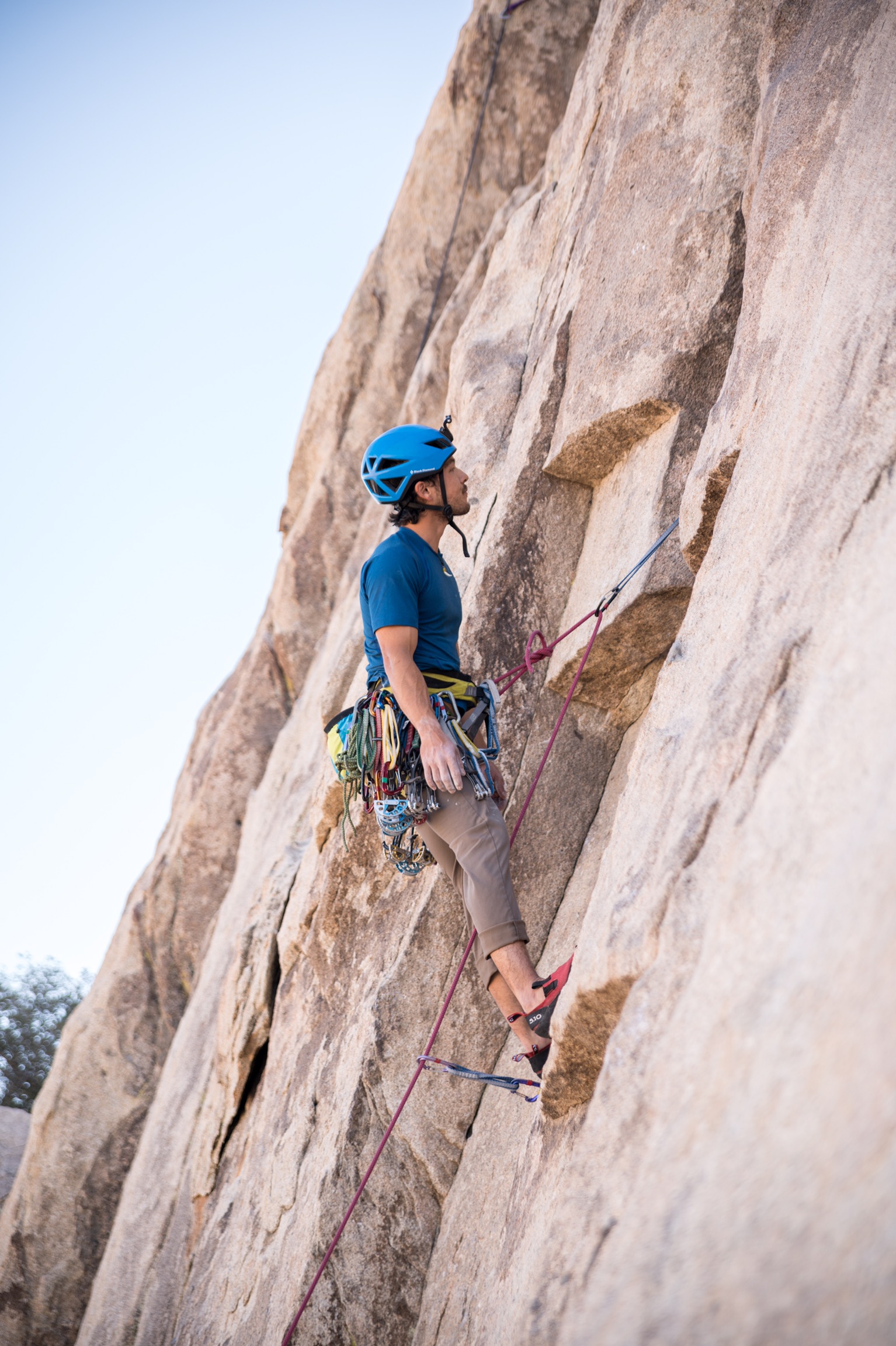 A man wearing a helmet and carrying climbing gear around his waist, looks at a massive rock face, as he is held to it by a rope.