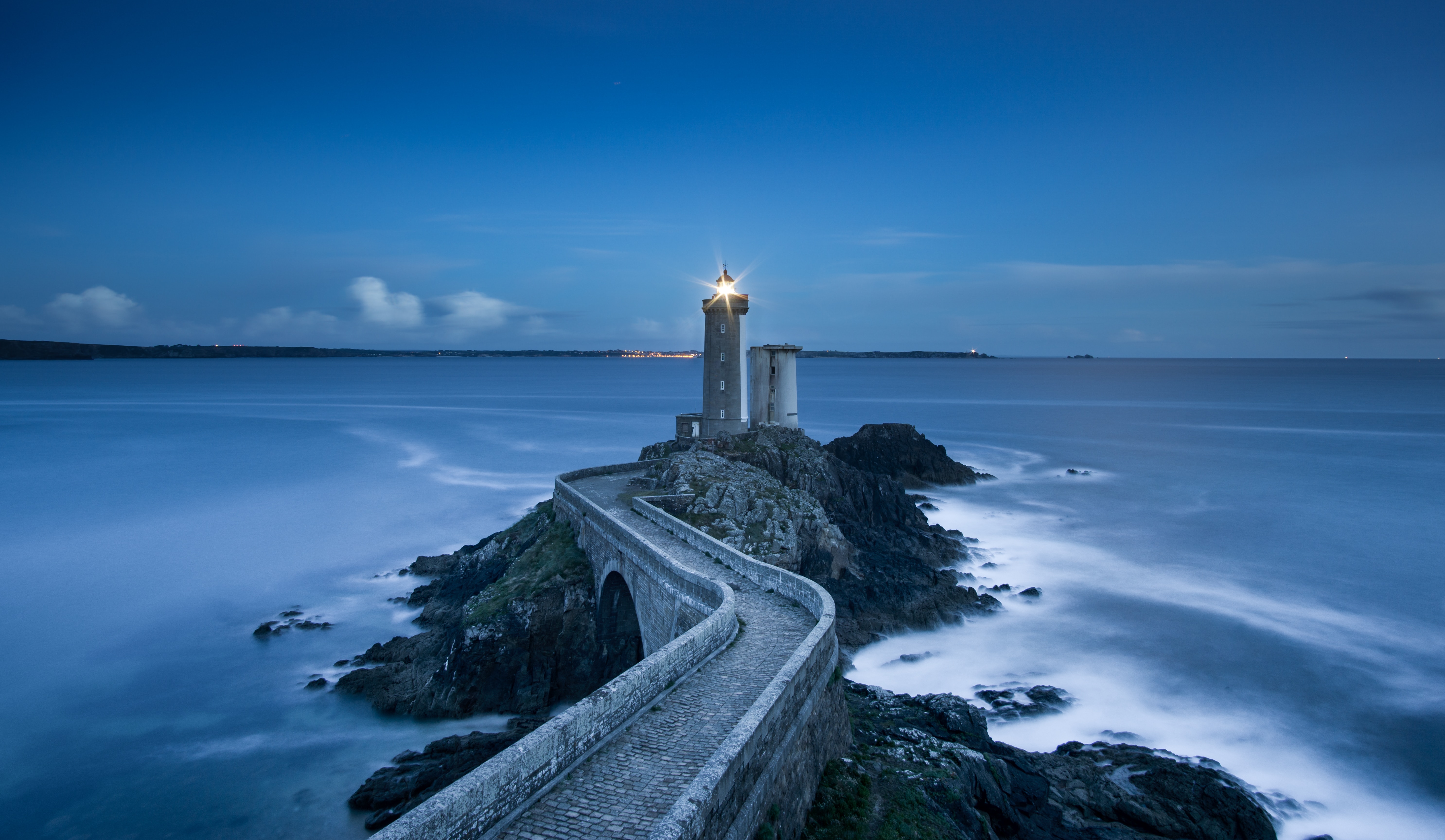 A winding stone path on a rocky outcrop leads to a small lighthouse. The light is shining in the darkness of blue hour.